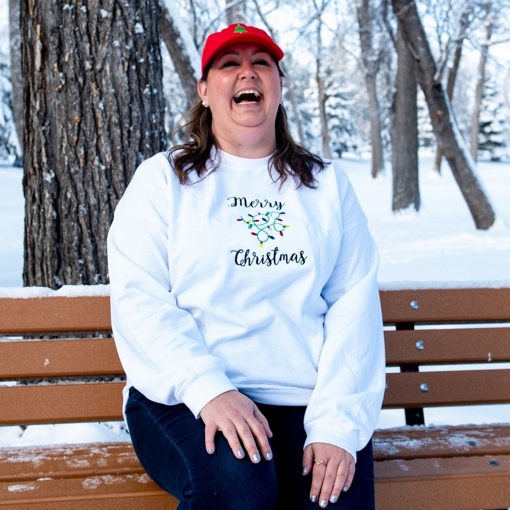 Female sitting on a bench wearing a white sweatshirt embroidered with Merry Christmas in the front - DSY Lifestyle