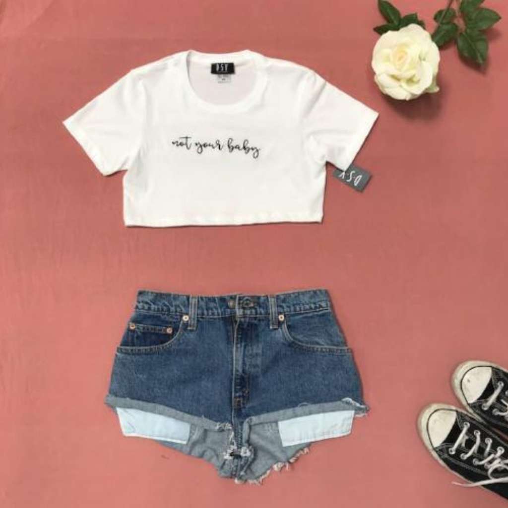 Flat lay of white underboob crop top with not your baby printed in black on the front paired with jean shorts and sneakers - DSY Lifestyle