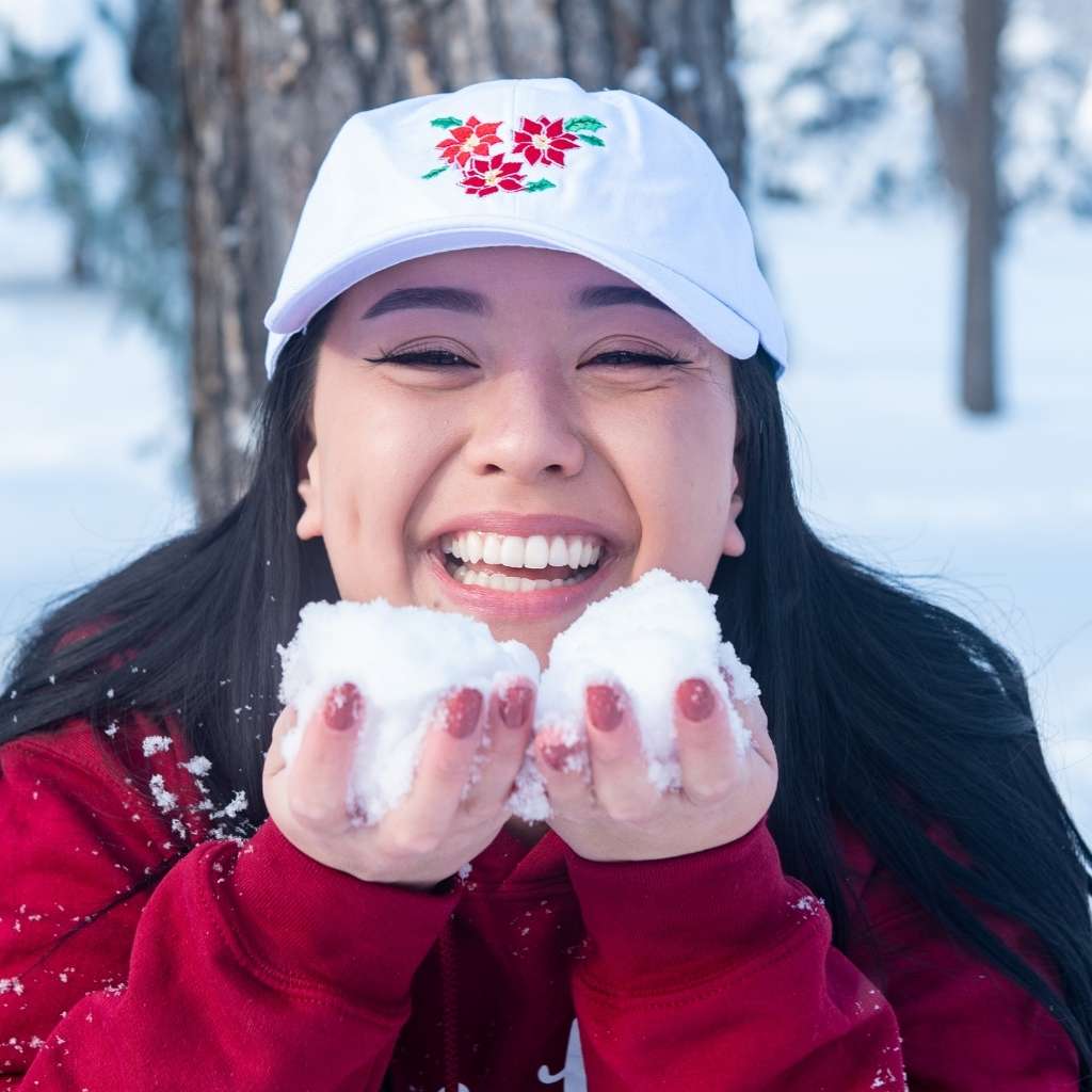 Female in the snow wearing a white baseball hat embroidered with poinsettias - DSY Lifestyle