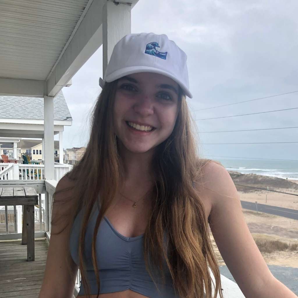 A woman wearing a white baseball cap with an embroidered wave design on the front, standing on a deck with an ocean view in the background. DSy Lifestyle